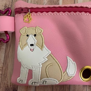 Op de afbeelding: Roze ritszakje met een geborduurd Collie-hondontwerp in wit en bruin. Het zakje heeft een roze botvormige rits en een gouden pootbedel.