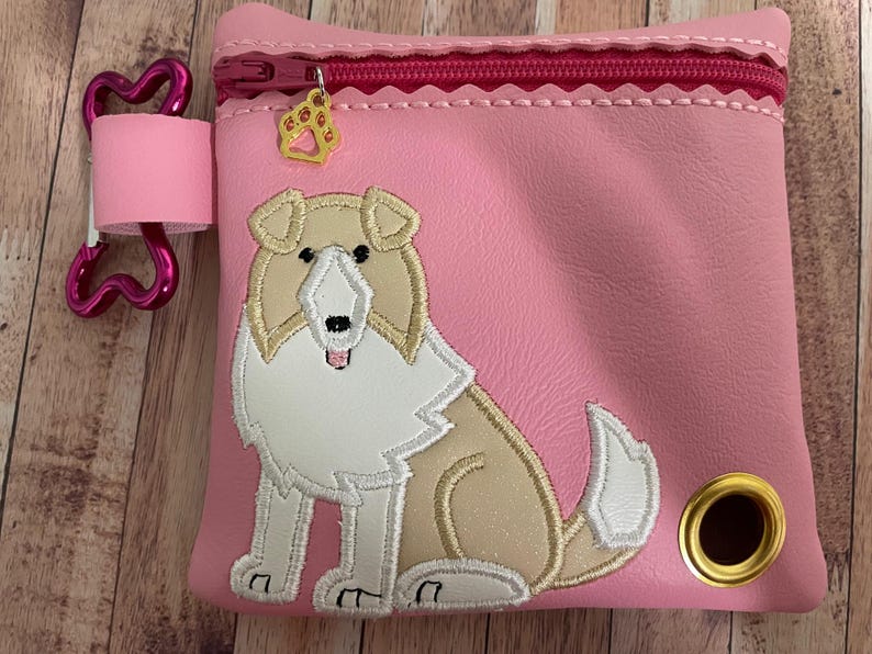 Op de afbeelding: Roze ritszakje met een geborduurd Collie-hondontwerp in wit en bruin. Het zakje heeft een roze botvormige rits en een gouden pootbedel.