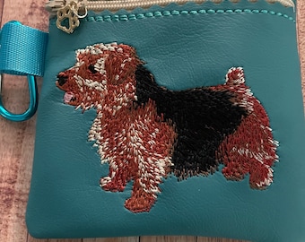 May include: Teal zippered pouch with a brown and black embroidered dog on the front. The pouch has a gold zipper pull with a paw print charm.