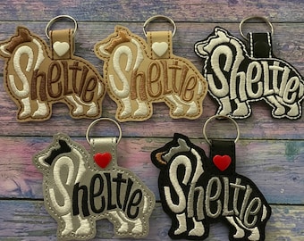 Op de afbeelding: Vijf verschillende sleutelhangers met een silhouet van een Shetland Sheepdog met het woord "Sheltie" erop geborduurd. De sleutelhangers zijn gemaakt van verschillende materialen, waaronder bruin, zilver en zwart stof. Elke sleutelhanger heeft een klein hartje eraan.