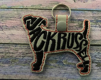 May include: A Jack Russell Terrier keychain in the shape of a dog. The keychain is gray with black embroidered text and a red stitched outline. It has a silver keyring and a gray snap closure.