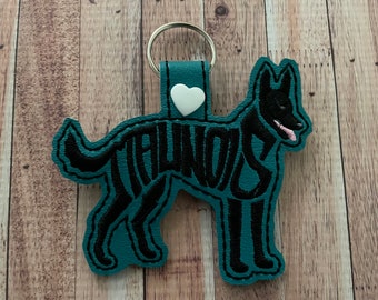 May include: Teal blue leather keychain shaped like a dog with the word "TAIWAN" spelled out in black embroidery. A white heart is attached to the top of the keychain.