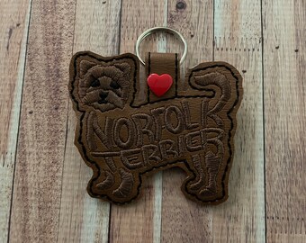 May include: Brown leather keychain with an embroidered Norfolk Terrier silhouette and the text "I love Norfolk Terrier". A red heart is sewn on the keychain.