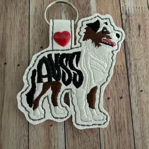 May include: A white and black embroidered keychain in the shape of an Australian Shepherd dog. The keychain has the word "AUSS" in black, a red heart, and a silver key ring.