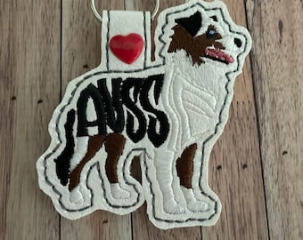 May include: A white and black embroidered keychain in the shape of an Australian Shepherd dog. The keychain has the word "AUSS" in black, a red heart, and a silver key ring.
