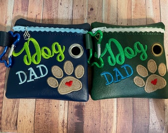 May include: Two small zippered pouches with a dog paw print and the text "Dog Dad" embroidered on them. One pouch is navy blue with green and light blue embroidery. The other pouch is dark green with green and light blue embroidery.