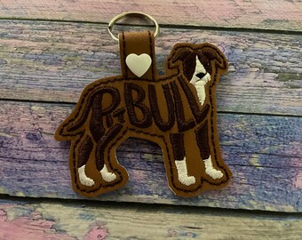 May include: Brown leather key chain with a white heart charm and an embroidered image of a brown and white pit bull with the word "P-BULL" spelled out in the dog's silhouette.