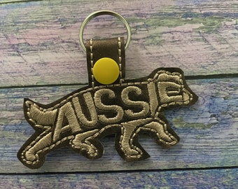 May include: A brown leather keychain with a silver key ring and a yellow button. The keychain is shaped like a dog and has the word "Aussie" embroidered on it in silver thread.