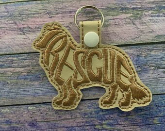 May include: A tan leather keychain with a gold embroidered silhouette of a dog. The dog's silhouette is filled with the word "RESCUE" in gold embroidery.