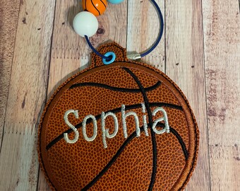 May include: A brown basketball-shaped ornament with the name "Sophia" embroidered in white on it. The ornament has a blue cord with white, orange, and blue beads attached to it.