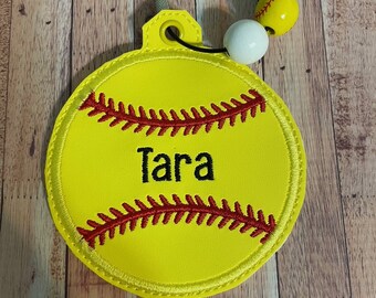 May include: Yellow softball keychain with red stitching and the name "Tara" embroidered in black. The keychain has a black cord with three beads: one white, one blue, and one yellow with red stitching.