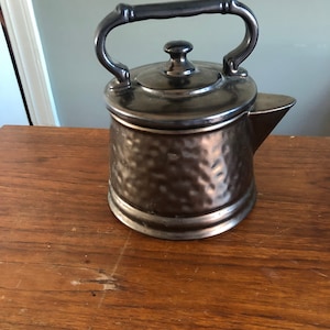 May include: A brown ceramic teapot cookie jar with a black handle and lid. The jar has a hammered metal finish.