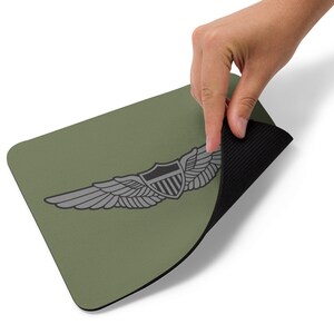 Pode incluir: Um tapete de rato verde com um emblema cinza de asas de piloto da Força Aérea dos Estados Unidos. O tapete de rato tem um suporte de borracha preto.