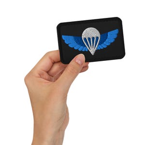 May include: Black embroidered patch featuring a white parachute design with blue wings. The patch is rectangular with a black border. The patch is held in a hand.