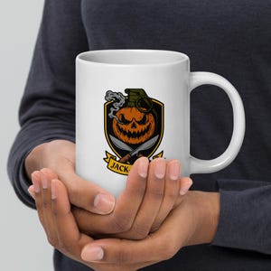 May include: White ceramic mug with a Halloween design. The design features a grinning jack-o'-lantern with a grenade top, crossed knives, and the word "JACK-" on a banner. The mug is held by a person.