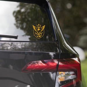 May include: A gold and black emblem sticker featuring an eagle with outstretched wings, a trident, and an anchor. The sticker is affixed to the rear window of a black vehicle. The emblem is a shield shape.