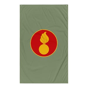 Puede incluir: Bandera verde oliva con un emblema central. El emblema es una forma de bomba amarilla con una llama amarilla encima, dentro de un círculo rojo. La bandera es rectangular con ojales en la parte superior para colgar.