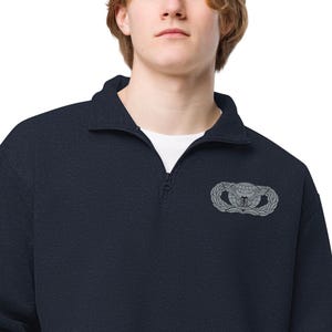 Könnte beinhalten: Marineblauer Pullover mit Viertelreißverschluss und weißem Unterhemd. Der Pullover hat ein silbernes Emblem auf der Brust, das aus einem Globus, Flügeln und einem Kranz besteht. Der Kragen ist im Fold-Over-Stil.