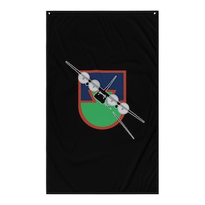 Puede incluir: Una bandera negra con un escudo verde, rojo y azul que presenta la silueta de un avión con cuatro hélices. El avión vuela en diagonal a través del escudo.