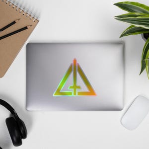 May include: A holographic sticker with a geometric design of a triangle, vertical line, and cross. The sticker has a green, yellow, and orange gradient. The sticker is on a silver laptop, with a notepad and pencils visible.