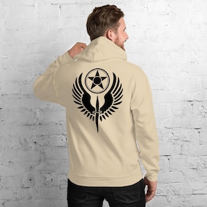 May include: A beige hoodie with a black graphic on the back. The graphic features a star inside a circle, wings, and a dagger. The hoodie has a hood and long sleeves. The person is wearing dark pants.