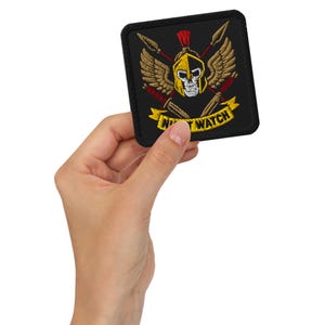 Pode incluir: Patch quadrado preto bordado com um capacete dourado e vermelho, uma caveira, asas e lanças cruzadas. O patch tem as palavras "NIGHT WATCH" em amarelo. O patch é segurado em uma mão.