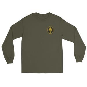 Pode incluir: Camiseta de manga comprida verde oliva com gola redonda. A camiseta apresenta um pequeno emblema circular no peito esquerdo. O emblema é um desenho de ponta de flecha dourada dentro de uma borda dourada em forma de corda. A camiseta é feita de material macio e confortável.