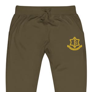 May include: Olive green sweatpants with a drawstring waist and side pockets. A gold emblem with text is embroidered on the right leg. The pants are made of a soft, comfortable material.