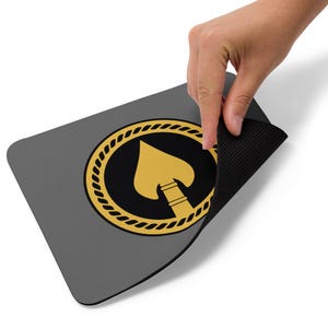 May include: A gray mouse pad with a black and gold design. The design features a stylized spearhead inside a circle with a rope border.