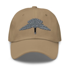 May include: A tan baseball cap with a detailed embroidered emblem featuring wings, a sword, and a cloud-like design. The cap has a curved brim and a classic six-panel construction, suitable for casual wear.