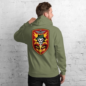 May include: An olive green hoodie with a large emblem on the back. The emblem includes a skull, wings, and the text "MAC SOG" within a shield design. The hoodie has a drawstring hood and long sleeves.