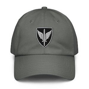 May include: A charcoal gray baseball cap featuring an embroidered patch on the front. The patch displays a black shield shape containing a white winged sword emblem. The cap has a curved brim, a button on top, and stitched eyelets for ventilation.