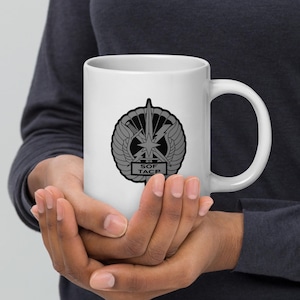 Könnte beinhalten: Weiße Keramik-Tasse mit einem grauen Emblem mit einem Stern, Flügeln und dem Text "SOF TACP". Die Tasse hat einen großen Henkel. Das Design ist mittig auf der Tasse und wird mit beiden Händen gehalten.