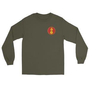 May include: Olive green long-sleeve shirt with a small, circular emblem on the upper left chest. The emblem features a red circle with a yellow border, containing a yellow flame and a yellow sphere.