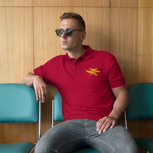 May include: A red short-sleeved polo shirt featuring a yellow embroidered emblem on the left chest. The emblem depicts a skull with wings and crossed swords. The shirt has a classic collar with a button placket and ribbed sleeve cuffs.