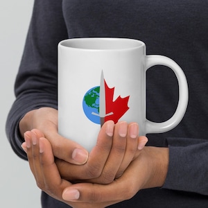 May include: White ceramic mug featuring a red maple leaf and a silver knife piercing a blue globe. The knife is positioned vertically, with the blade facing upwards.
