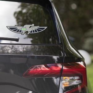 May include: A black car with a silver and green cat-shaped sticker on the rear window. The sticker has wings and a green face.