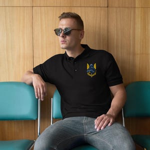 Op de afbeelding: Zwarte poloshirt met een klein geborduurd embleem op de linkerborst. Het embleem toont een donkere figuur met gele vleugels en tekst. De shirt heeft een klassieke kraag en een knoopsluiting.
