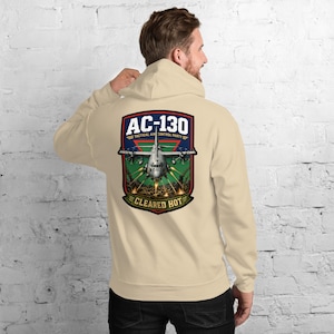 May include: A beige hoodie featuring a large graphic on the back. The graphic includes the text "AC-130 TACTICAL AIR CONTROL PARTY" above an illustration of a military aircraft, with the words "CLEARED HOT" below.