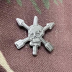 Op de afbeelding: Een zilverkleurige metalen pin met een schedel met gekruiste pijlen en een dolk. De schedel heeft gedetailleerde gelaatstrekken en de pijlen zijn voorzien van veren. De pin is waarschijnlijk een militair of tactisch embleem.