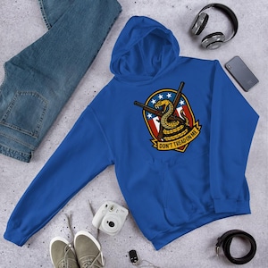 May include: A royal blue hoodie featuring a graphic of a coiled snake with crossed rifles, a red, white, and blue shield, and the text "DON'T TREAD ON ME." The hoodie is paired with blue jeans, sneakers, and accessories.