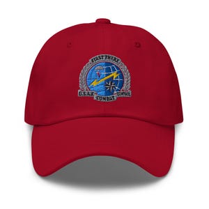 May include: A red baseball cap with a curved brim. The cap features an embroidered patch with the text "FIRST THERE" above a globe design with a yellow arrow and a star. The text "U.S.A.F. COMBAT CONTROL" is also embroidered on the patch.