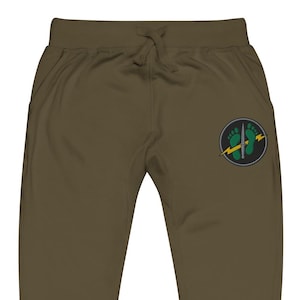 May include: Olive green sweatpants featuring an elastic waistband with a drawstring. A circular patch on the right leg displays a graphic of two green footprints, a vertical silver dagger, and a yellow lightning bolt against a dark background.