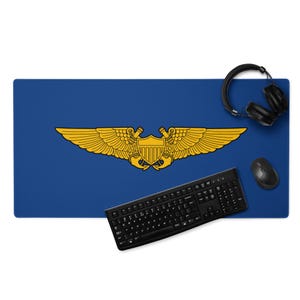 Könnte beinhalten: Blaues Schreibtisch-Pad mit einem goldenen Emblem aus Flügeln und einem Schild. Eine schwarze Tastatur, Maus und Kopfhörer liegen auf der Matte. Das Schreibtisch-Pad ist rechteckig.