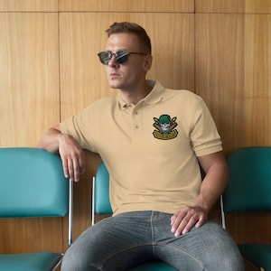 May include: A pale beige polo shirt featuring a skull and baseball bats emblem. The emblem includes a green helmet and the text "DEATH BOY WORKS". The shirt has a traditional collar and short sleeves.