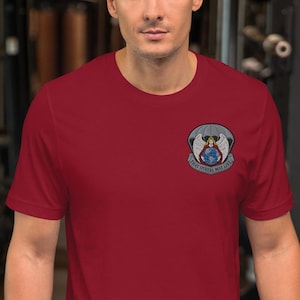 Può includere: T-shirt bordeaux con una toppa ricamata raffigurante un globo, ali e la scritta "THAT OTHERS MAY LIVE". La toppa è centrata sul petto. La maglietta è realizzata in un tessuto morbido.