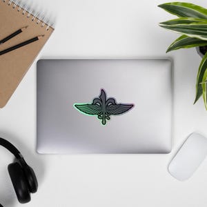 May include: A sticker featuring a stylized emblem with a fleur-de-lis design and wings, with a gradient of green, pink, and purple. The sticker is on a laptop, surrounded by office supplies and a plant.