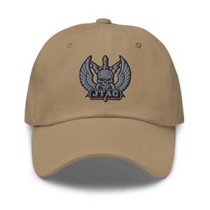 May include: A tan baseball cap with a detailed embroidered emblem. The emblem features a skull with wings, a sword, and the text "JTAC" in red. The cap has a curved brim and an adjustable strap.