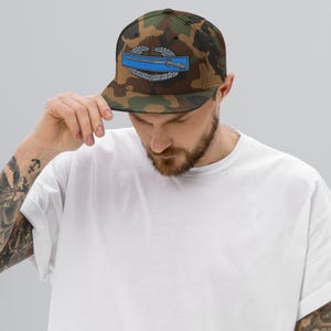 Può includere: Cappellino da baseball mimetico con un emblema blu e argento con un design a fucile e corona d'alloro. Il cappellino ha una visiera piatta e una vestibilità strutturata. È un accessorio in stile militare.