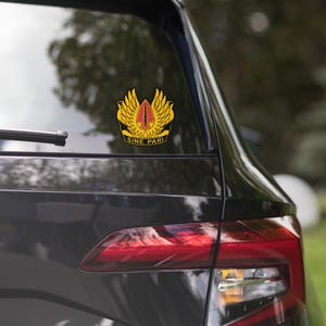 May include: A black car with a yellow and red decal on the rear window. The decal features a winged symbol with a sword and the words "Sine Pari".